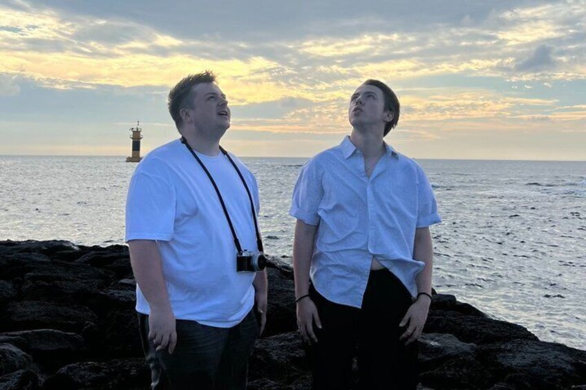 Two people standing on a rocky shore with a lighthouse in the background. 5