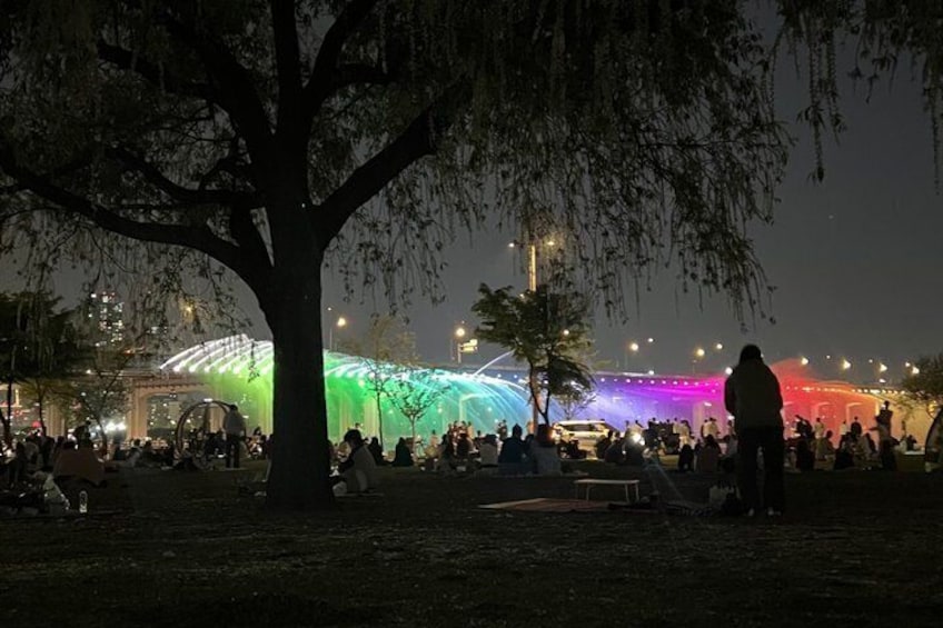 A nighttime outdoor gathering with a large tree in the foreground and a brightly lit structure in the background. 5