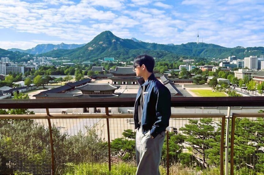 A person standing on a balcony overlooking a traditional Korean palace, with mountains and modern buildings in the background. 2
