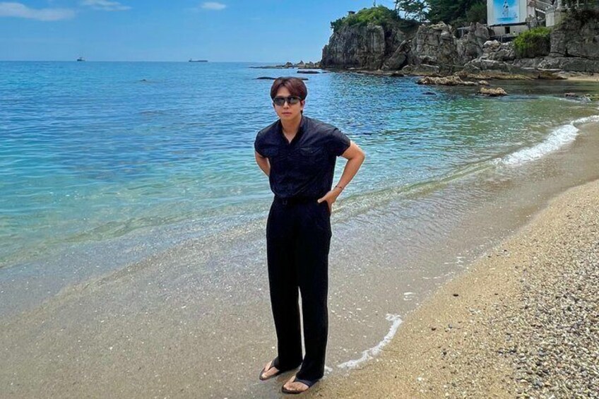 A person standing on a pebbly beach with clear blue water and rocky cliffs in the background. 3
