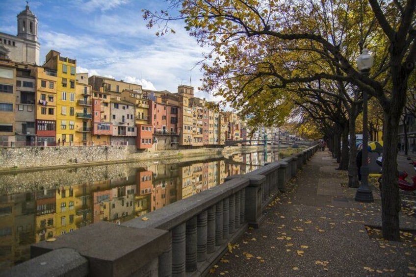 A riverside walkway with colorful buildings, a bridge, and autumn leaves. 3