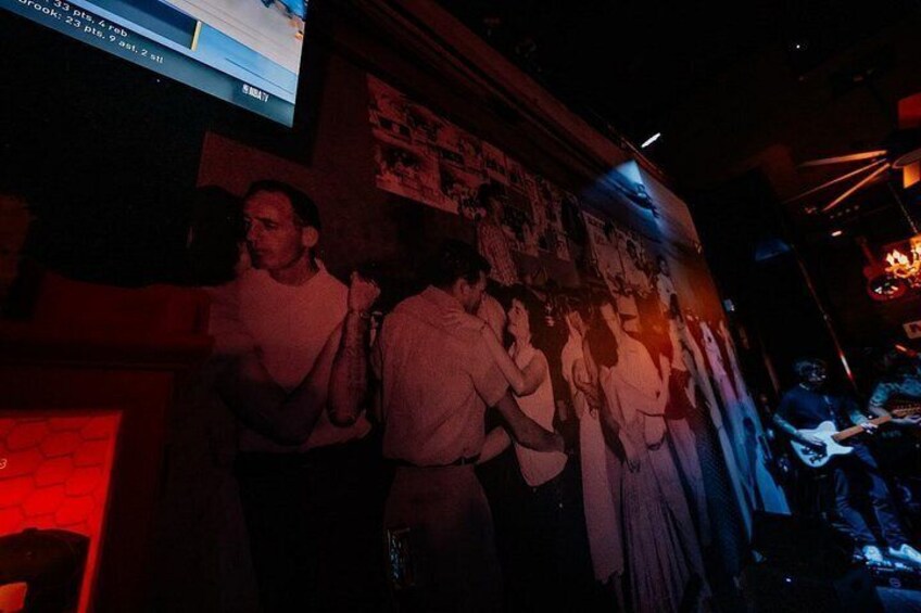 A dimly lit room with a mural on the wall, a person playing a guitar, and a television screen displaying text. 2