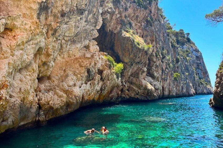 A rocky coastline with clear turquoise water and two people swimming. 4