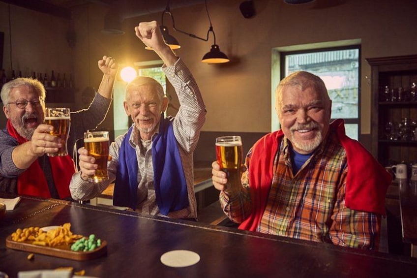 Four people at a bar, holding beer glasses, with a tray of snacks in the foreground. 4