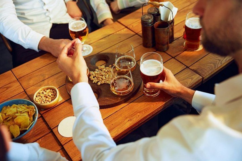 A wooden table with people seated around it, holding glasses of beer, and a bowl of peanuts. 3