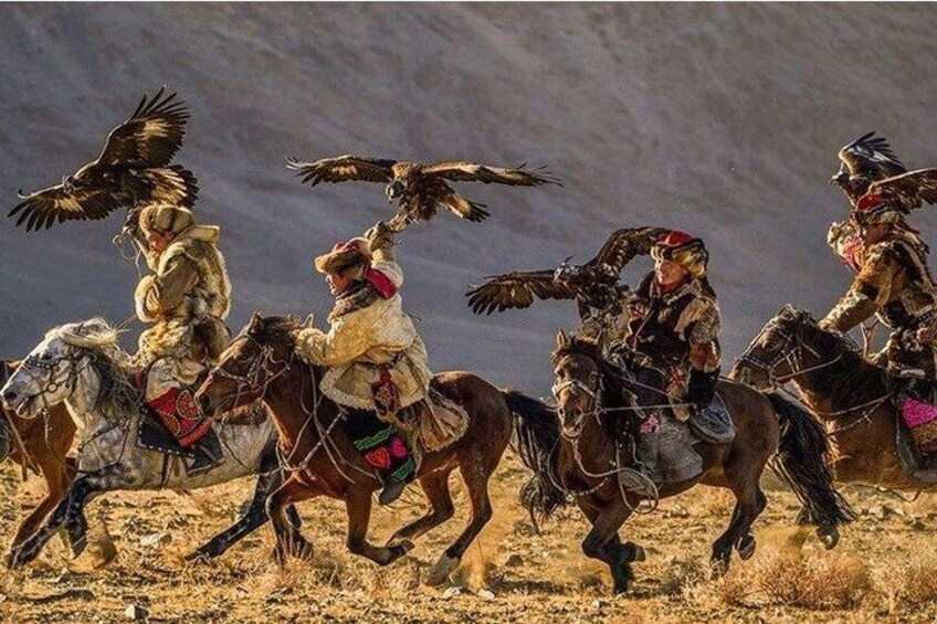 A group of mounted individuals in traditional attire, riding horses across a dry, grassy terrain with mountains in the background. 1