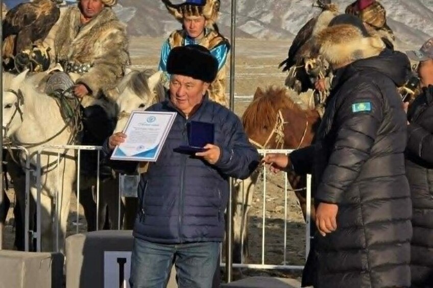 A person holding a certificate, standing in front of a barrier with horses and people in traditional attire. 4