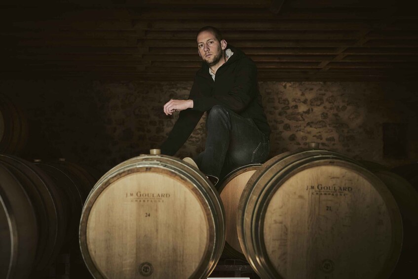A person sitting on a barrel in a cellar with wooden walls and barrels labeled 'J.M. GOUILLARD.' 2