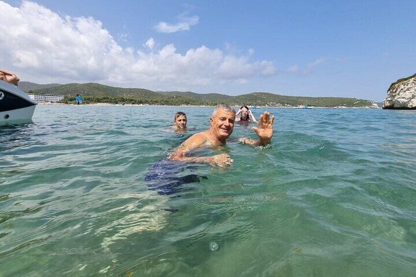 People swimming in clear water with a boat and rocky coastline in the background. 3