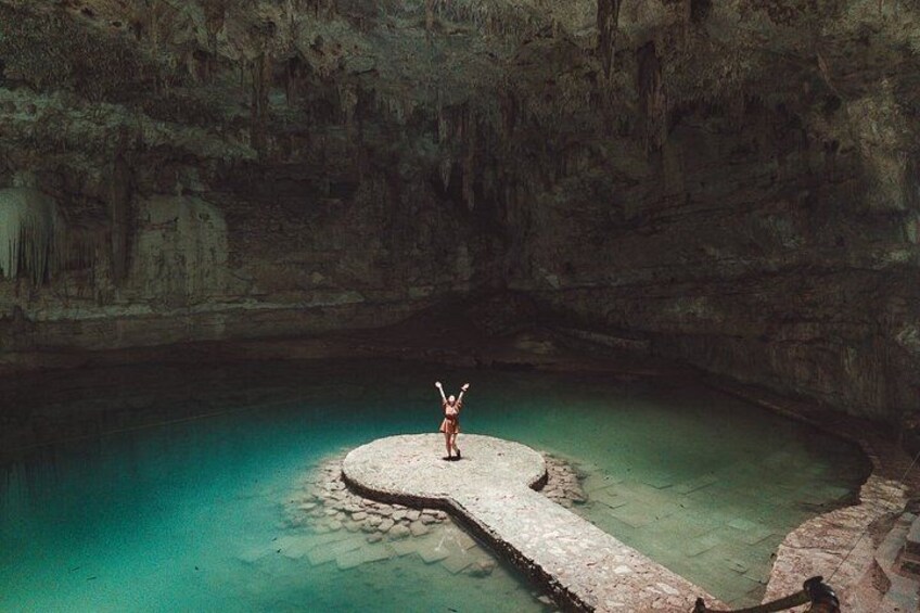 A person standing on a circular stone platform in a cave with clear turquoise water. 1