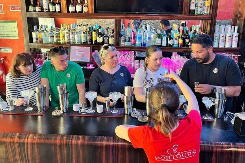 A bar with people seated at tables, a bartender preparing drinks, and a variety of bottles and glasses on shelves. 1