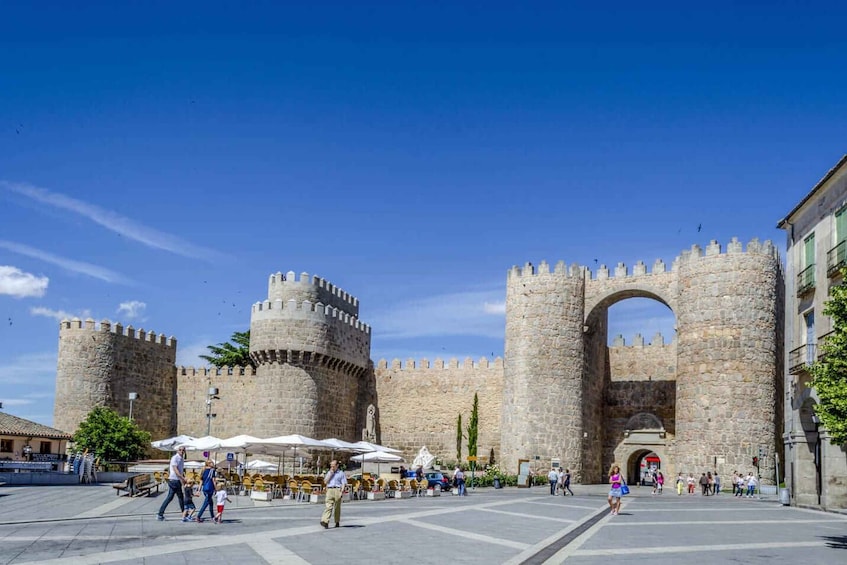 A historic stone fortress with a large archway, surrounded by a square plaza with cafes and pedestrians. 1
