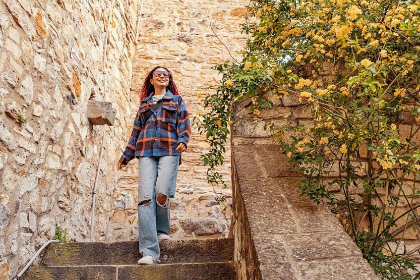 A person standing on stone steps in a narrow alleyway with a stone wall and a flowering bush. 3