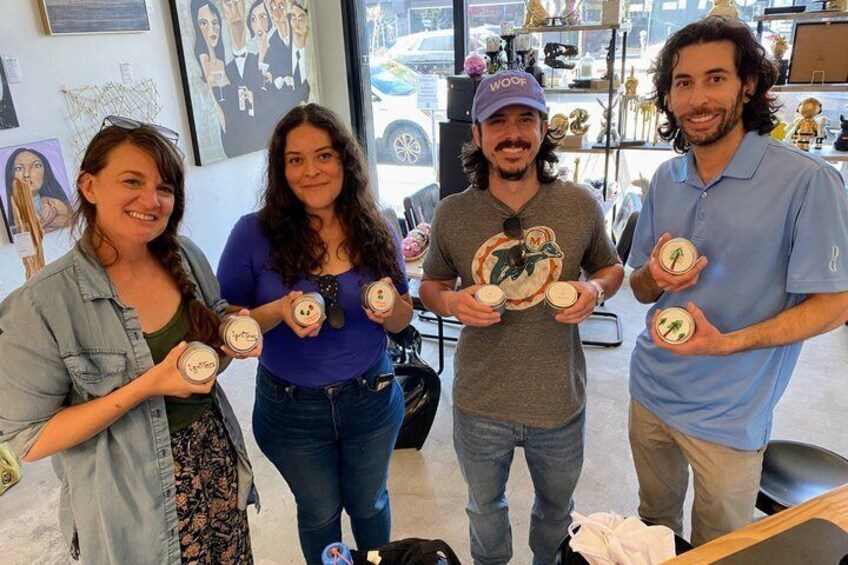Four people holding small jars with labels, standing indoors with artwork on the walls. 3