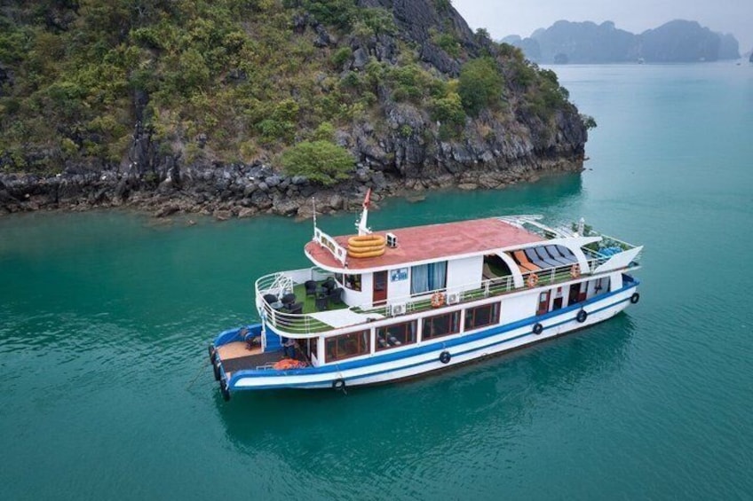 A large boat with a white and blue hull, a red roof, and multiple decks, anchored near a rocky coastline with clear turquoise water. 1