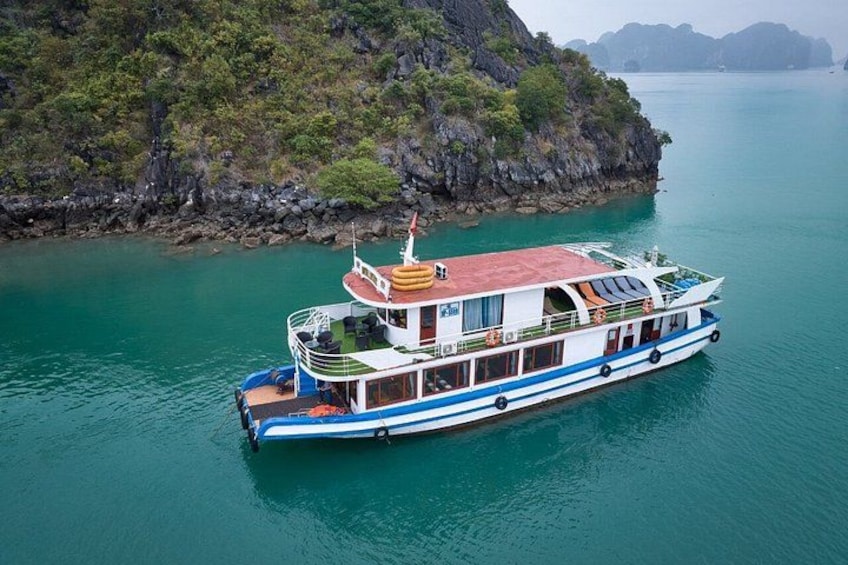 A large boat with multiple decks, white and blue color scheme, anchored near a rocky coastline with clear turquoise water. 1