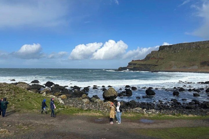 A rocky coastline with waves crashing, a clear sky, and people walking along the shore. 2
