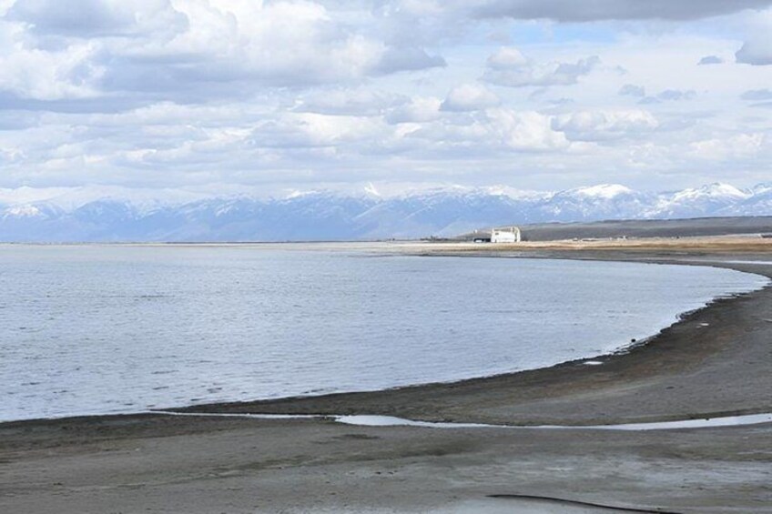A calm lake with a sandy shore, a small white building in the distance, and snow-capped mountains in the background. 5