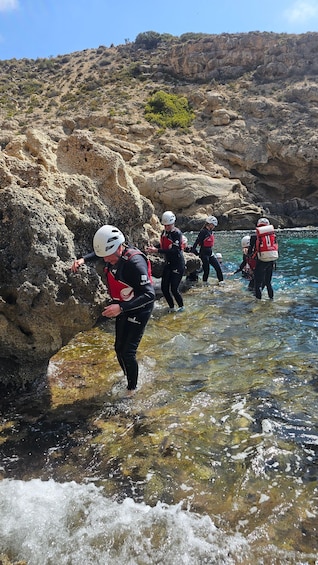 A group of people in wetsuits and helmets near rocky cliffs by a clear water body. 4