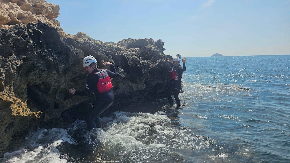 Two people climbing a rocky cliff by the sea, wearing helmets and life jackets. 2