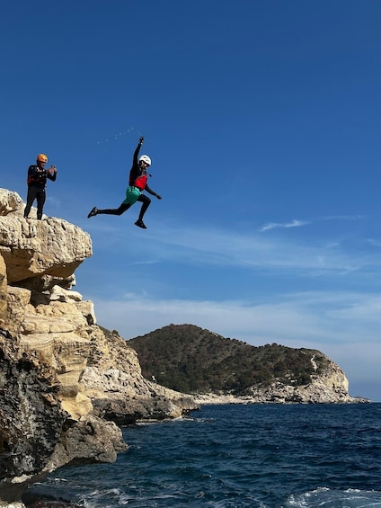 Two people jumping off a cliff into the sea, with rocky terrain and a distant hill in the background. 5