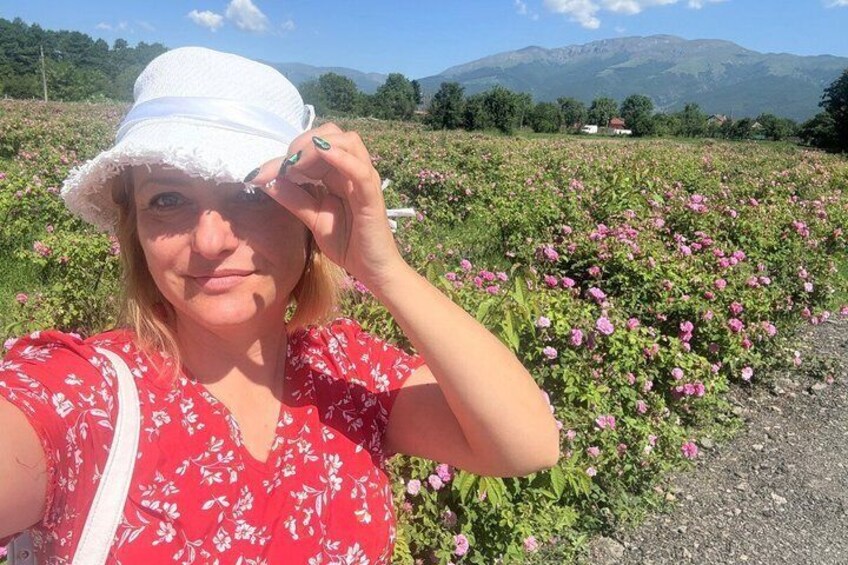 A person in a red floral dress and white hat taking a selfie in a field of pink flowers with mountains in the background. 1