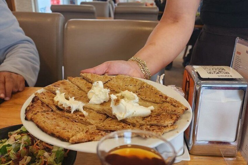 A person holding a plate with a large, round, layered pastry topped with whipped cream, a glass of a dark beverage, and a salt shaker on a table. 5