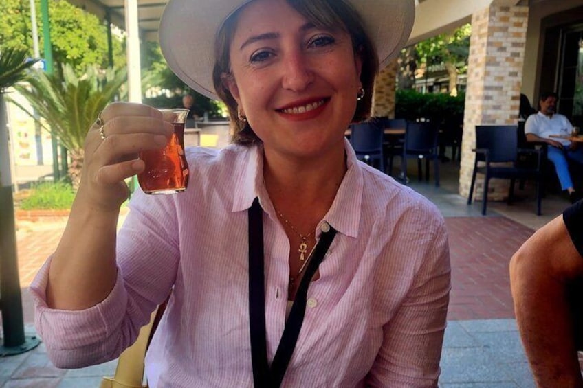 A person holding a glass of amber liquid, wearing a light pink shirt and a wide-brimmed hat, seated at an outdoor cafe with tables and chairs. 4