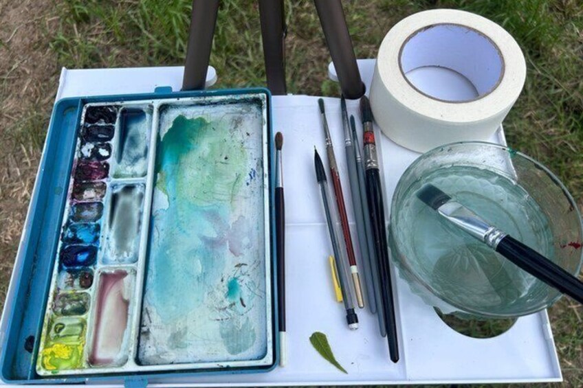 A watercolor palette, brushes, and a glass of water on a table outdoors. 4