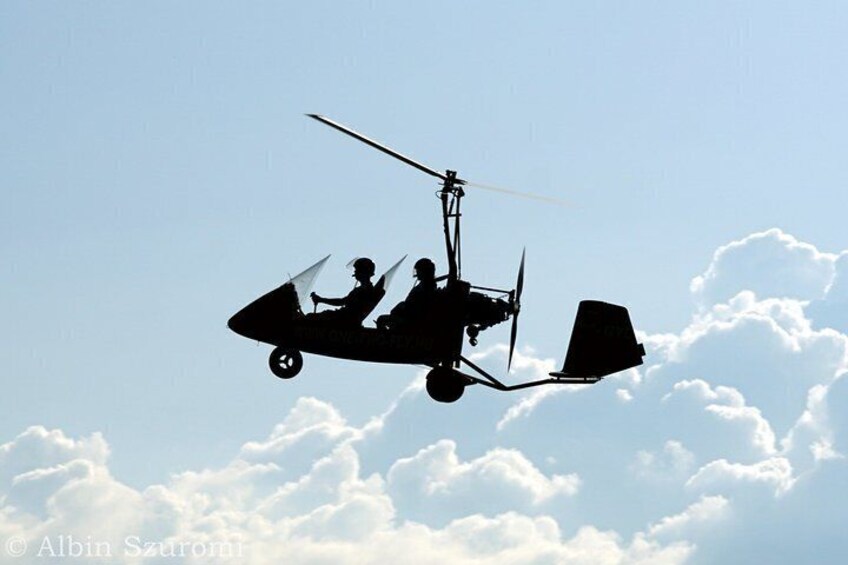 A helicopter with two people, one seated and one standing, flying above clouds. 2