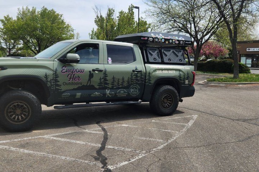 A green pickup truck with 'Empower Her Outdoors' branding parked in a parking lot. 2