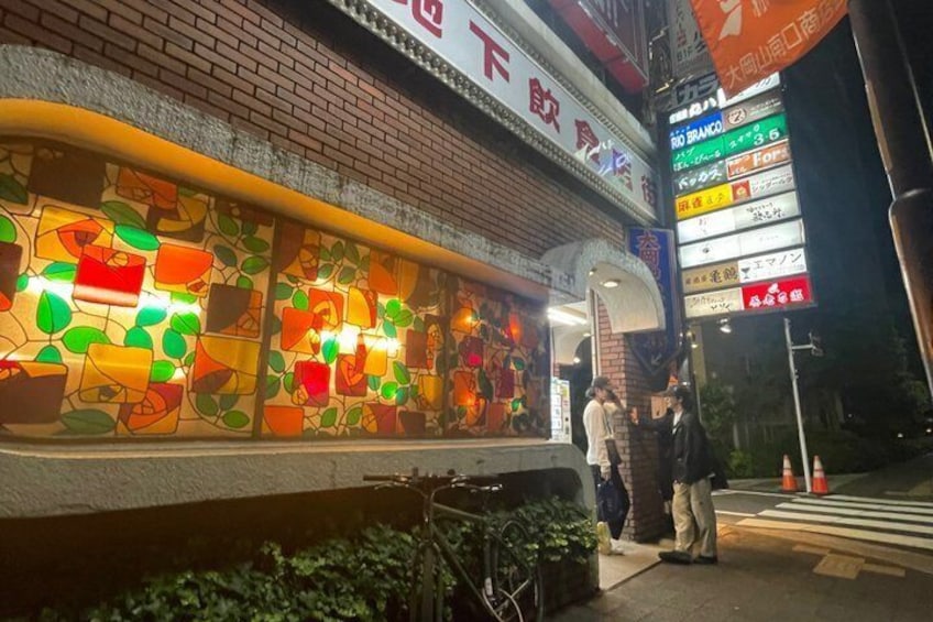 A brightly lit storefront with a colorful stained glass window and a signboard with multiple destinations. 5