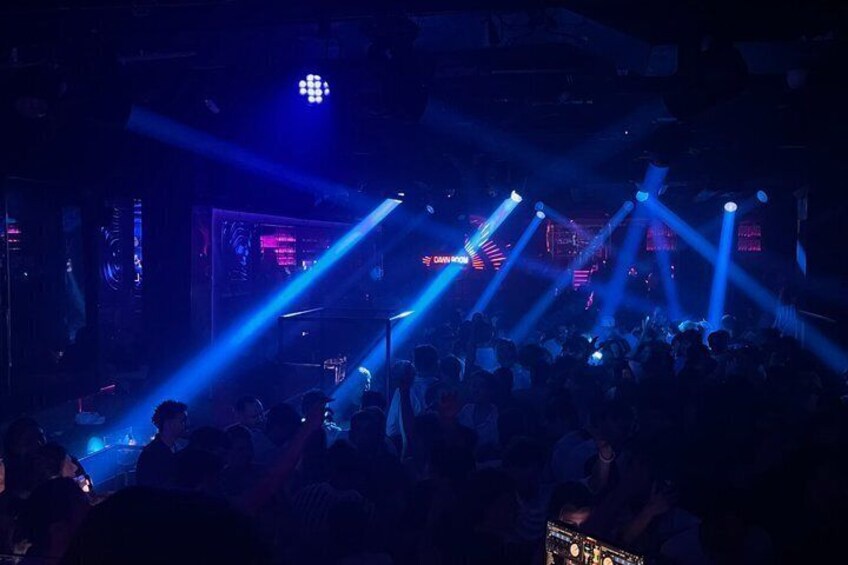 A crowded indoor venue with blue stage lighting and a DJ booth. 4