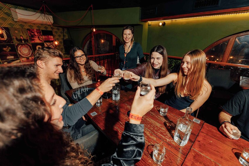 A group of people toasting with drinks around a wooden table in a dimly lit room with green walls and a vintage car in the background. 1