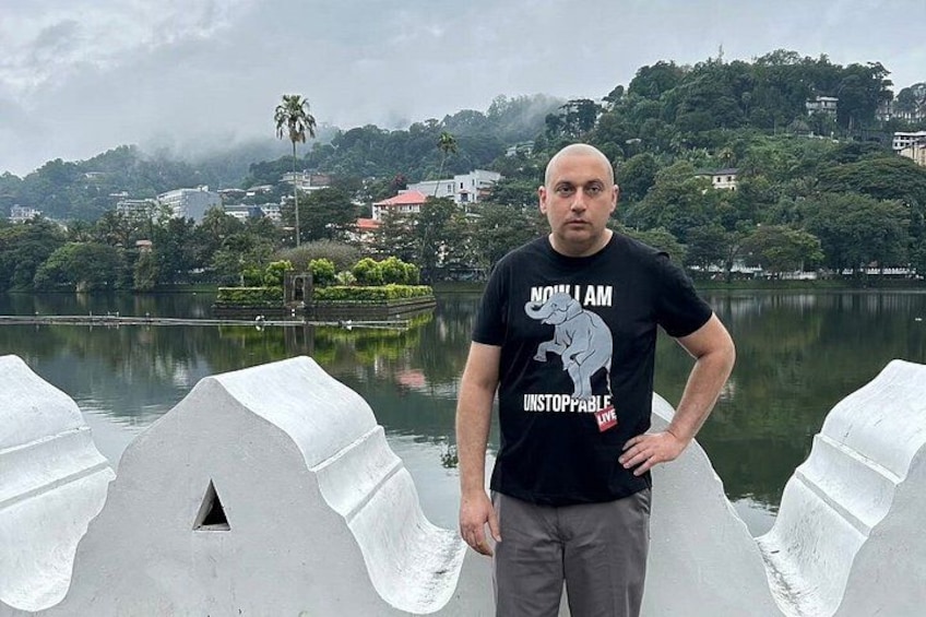 A man standing by a lake with white barriers, wearing a black t-shirt with a graphic and text, and a cloudy sky in the background. 4