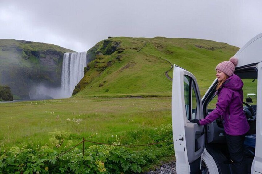 A person in a purple jacket and pink beanie standing next to a white vehicle with an open door, with a waterfall in the background. 2