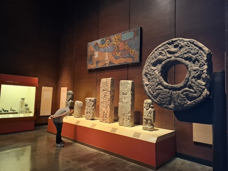 Intérieur d’un musée où sont exposées des sculptures en pierre. On aperçoit une grande pierre circulaire sur la droite, et une personne qui examine les sculptures. 5