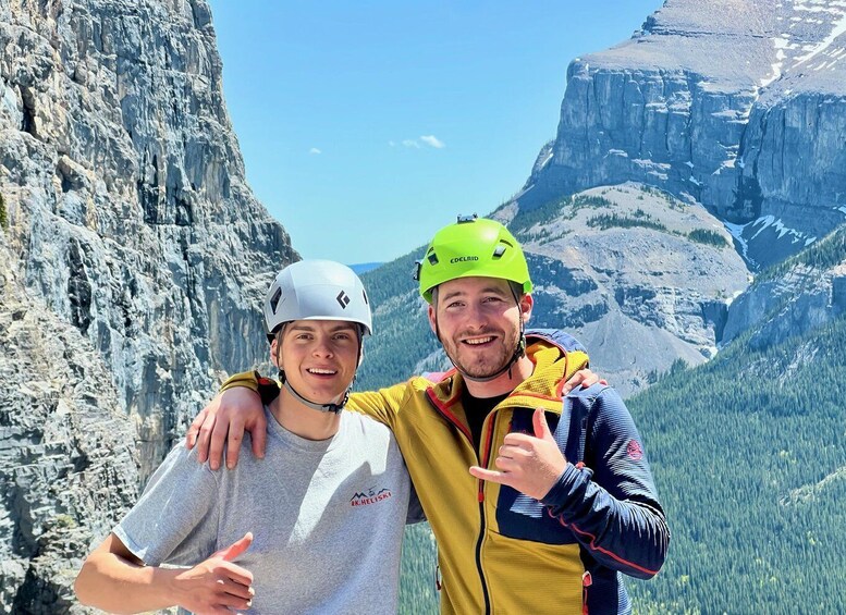 Two people wearing helmets, one in a grey shirt and the other in a yellow jacket, standing in front of a mountainous landscape with rocky cliffs and a clear sky. 2