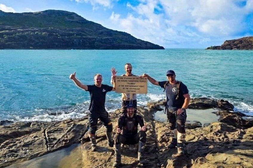 Four people standing on a rocky shoreline with a sign in the center. 4