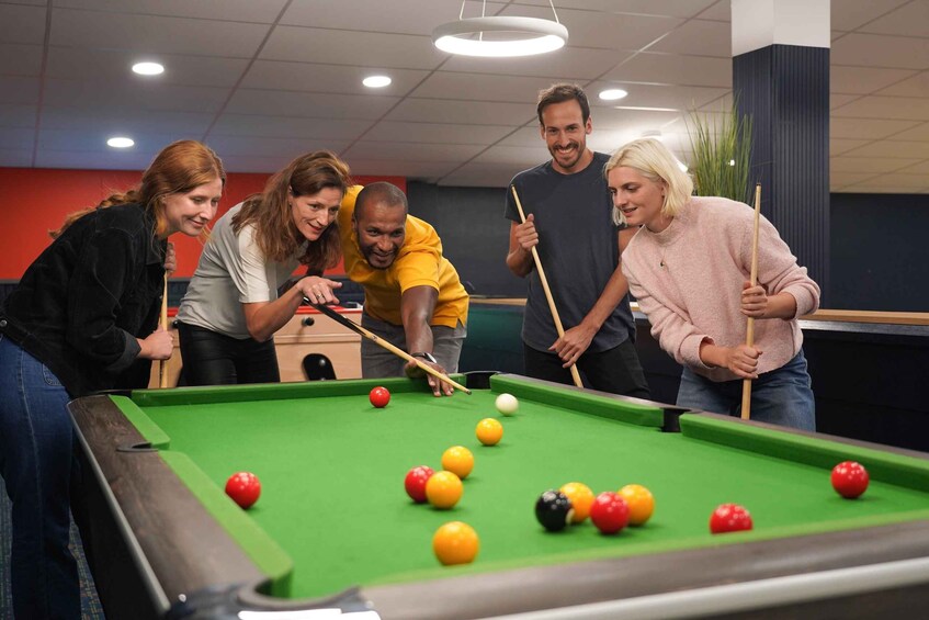 A group of people playing pool in a room with a green pool table, red and yellow balls, and a white ball. 5