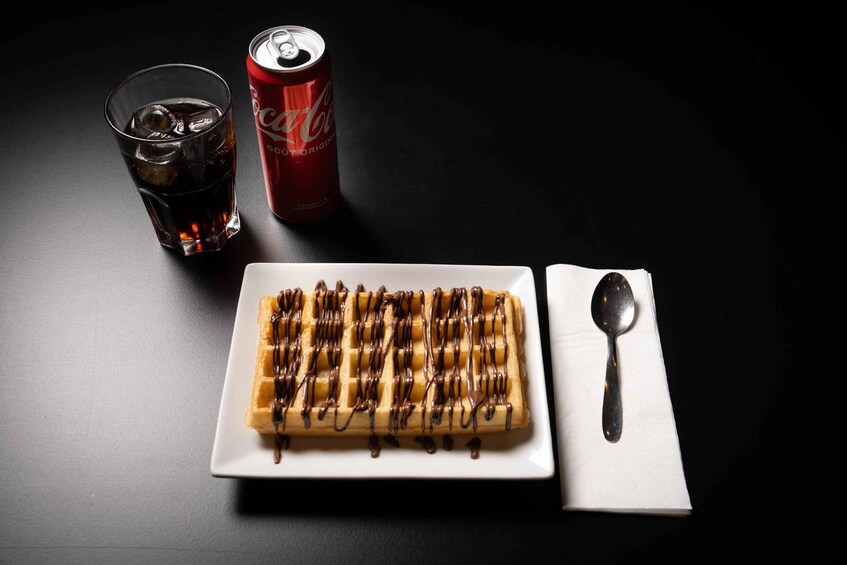A waffle with chocolate drizzle, a glass of cola, and a spoon on a table. 2