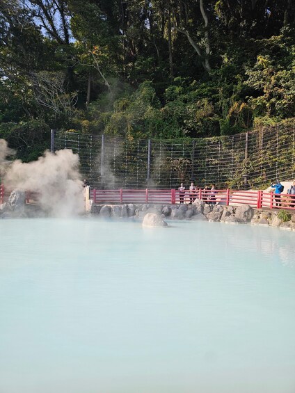A geothermal spa with steam rising from the water, surrounded by a red fence and trees. 2