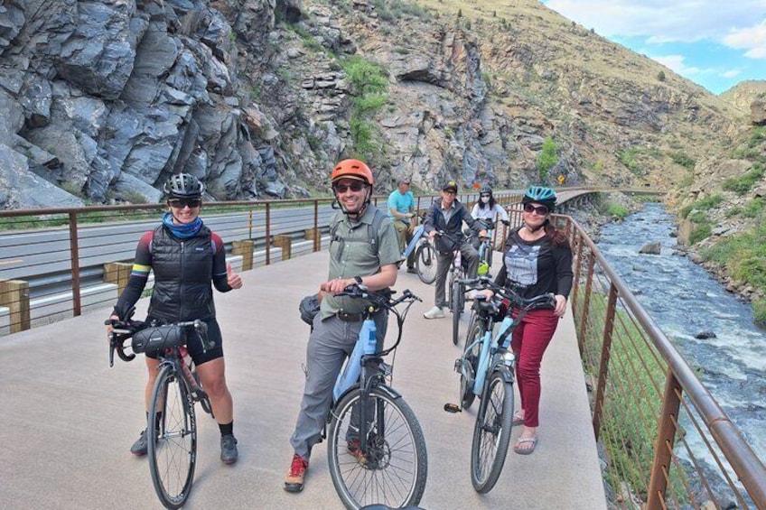 A group of cyclists on a paved bridge with a rocky riverbed and mountains in the background. 1