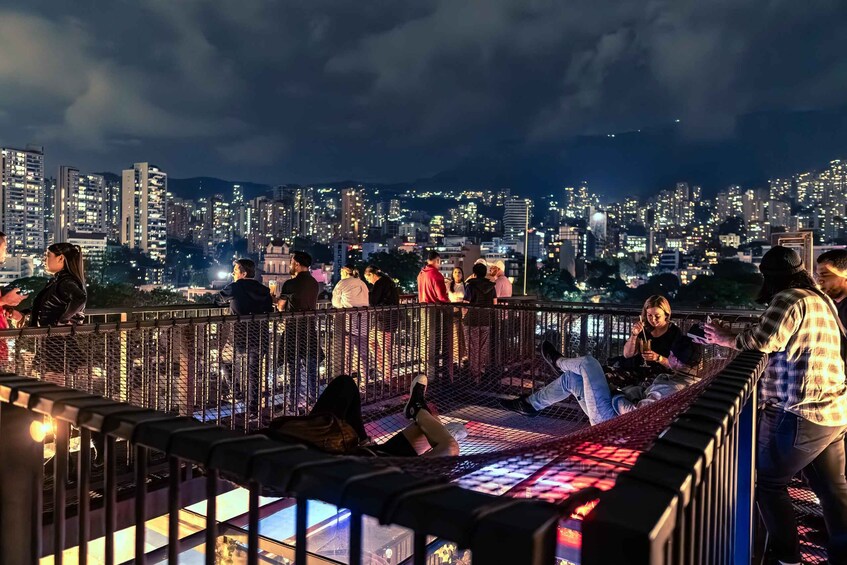 A rooftop bar with people sitting and standing, overlooking a cityscape at night. 1