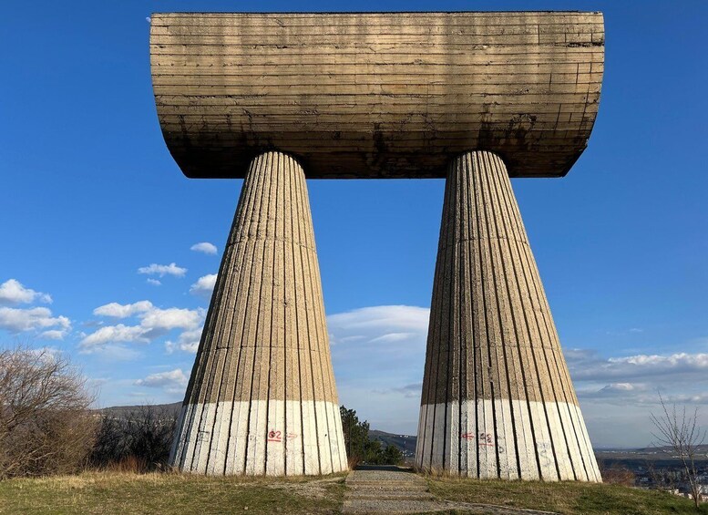 A large concrete structure with two vertical pillars supporting a horizontal beam, set against a clear blue sky. 3