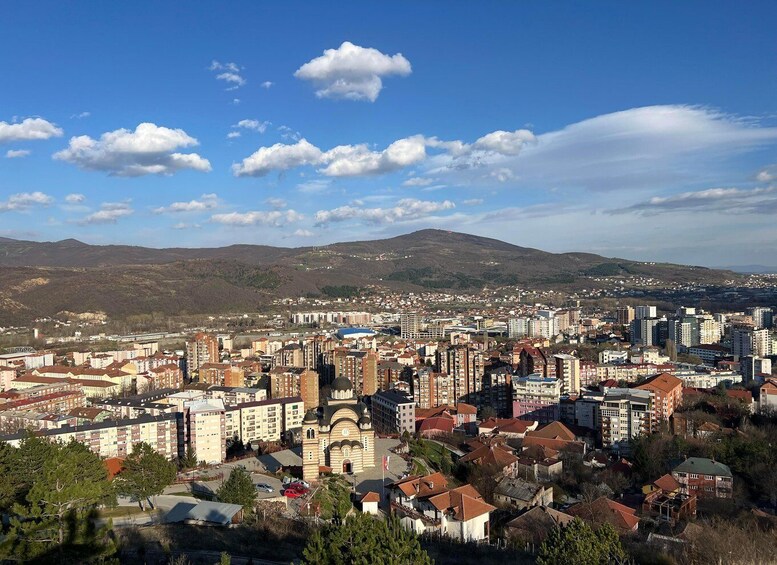 A cityscape with a mix of modern and traditional buildings, a prominent church, and a mountainous backdrop under a clear sky. 1