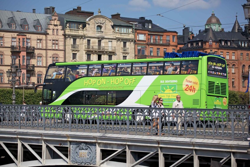 A green double-decker bus with 'HOP ON - HOP OFF' and '24h' text, traveling on a bridge. 5