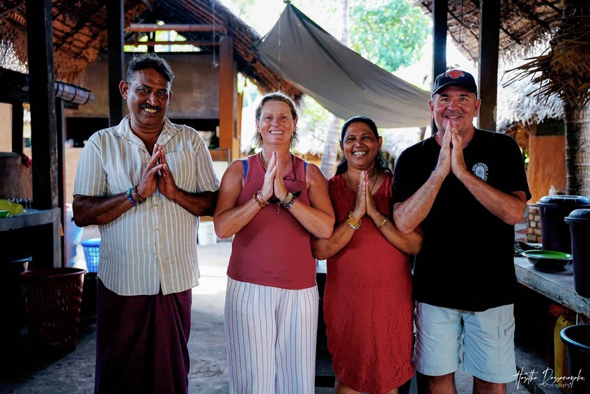 Four people standing in a covered outdoor area with a thatched roof, a table with containers, and a concrete floor. 1