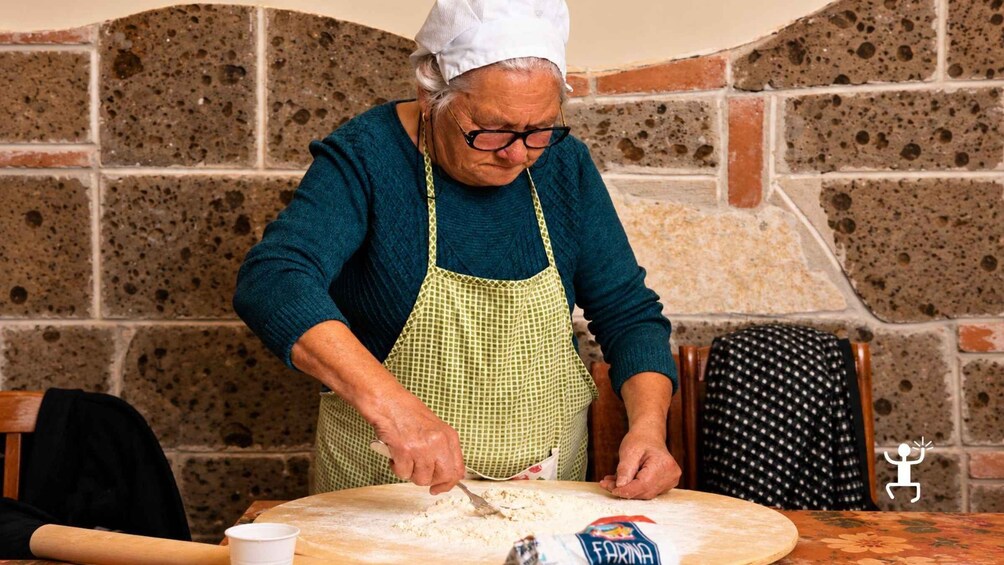 An elderly person wearing a chef's hat and apron, preparing food on a table with a rolling pin and a flour container. 2