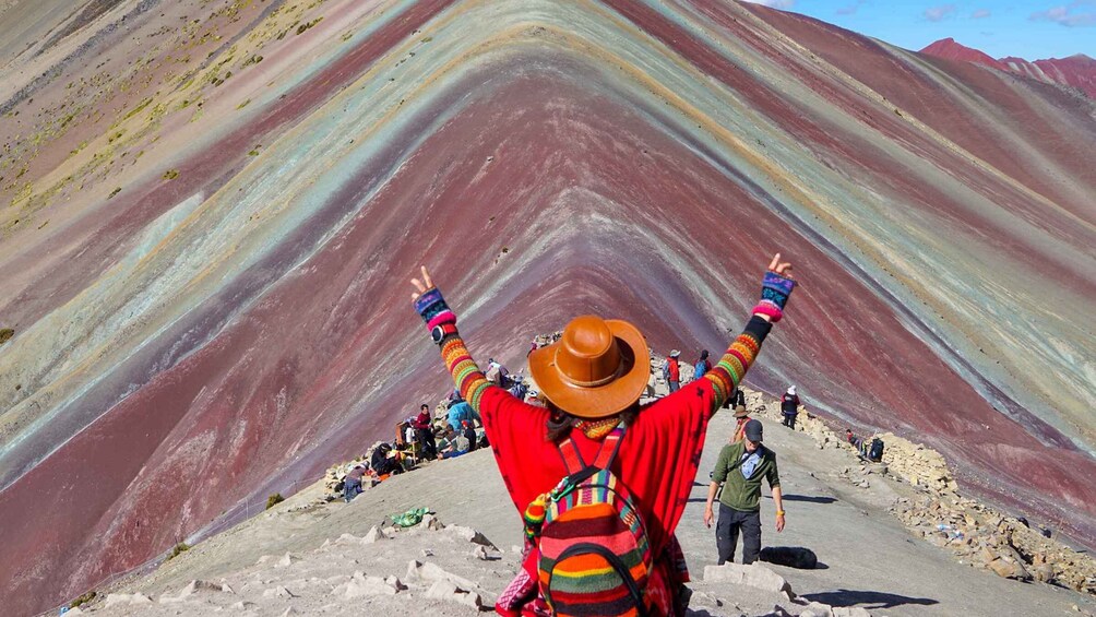 A person standing on a mountain ridge with arms raised, facing a colorful stratified rock formation. 3